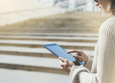 Hipster tourist texting message on tablet or technology mockup. Person using computer on building background close. Female hands holding gadget on blurred backdrop. Copy space for text, flareの写真素材