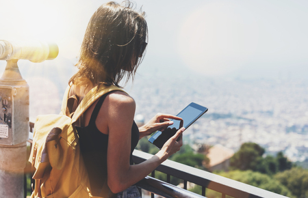 Hipster girl with backpack holding gadget in observation view. Tourist traveler on background panoramic city Barcelona, female hands using tablet and map, coin operated binoculars, sun flare, mockupの写真素材