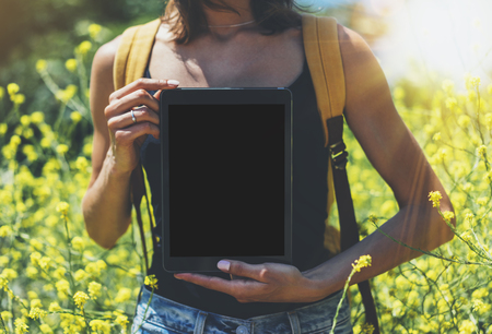 Hipster holding in hands tablet computer, mockup of blank empty screen. Girl traveler using gadget on sun flare and yellow flowers background close. Female photographer take view on cell cameraの写真素材