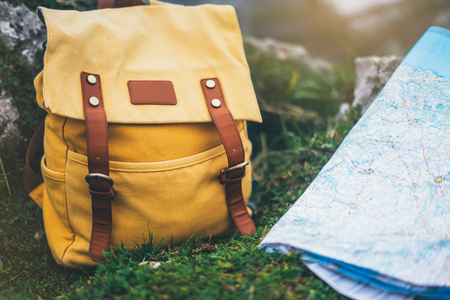 Hipster hiker tourist yellow backpack closeup and navigation map europe on background green grass nature, blurred panoramic landscape, traveler relax holiday concept, view planning way road in trip vacationの写真素材