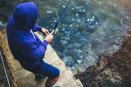 young man holds a fishing rod and catches fish in the nature on a sea background, hipster fisherman spends vacation on the blue ocean, active travel hobby fishing, rural outdoor sport tourism, top viewの写真素材