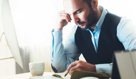 Bearded young businessman working on modern office. Consultant man thinking looking in monitor computer. Manager writes in notebook in coworking workplace, startup project concept in studio with coffeeの写真素材