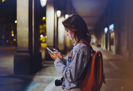 blogger hipster using in hands gadget mobile phone, woman with backpack pointing finger on blank screen smartphone on background bokeh light in night atmospheric city, mockup street, online wifi internet conceptの写真素材