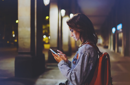 blogger hipster using in hands gadget mobile phone, woman with backpack pointing finger on blank screen smartphone on background bokeh light in night atmospheric city, mockup street, online wifi internet conceptの写真素材