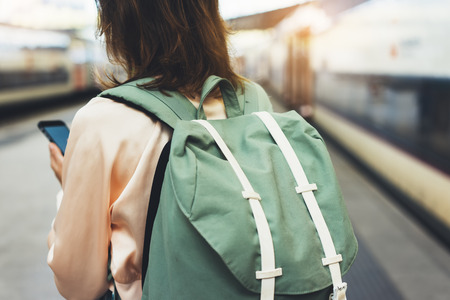 Enjoying travel. Young hipster woman waiting on the station platform with backpack on background electric train using smartphone. Tourist texting message and plan route of railway, railroad transport conceptの写真素材