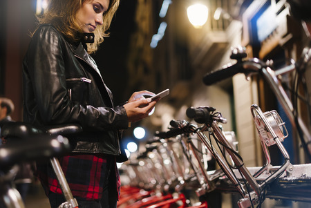 Young girl in black leather jacket using in hands screen smartphone on background bokeh light in night city street, hipster biking and riding to job by ecology  bicycle, lifestyle online wifi internet conceptの写真素材