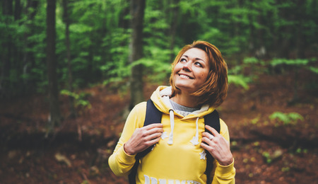 smiling tourist traveler with backpack into road at summer green forest, view back girl hiker in yellow hoody looking and enjoying the breath of fresh clean air in trip, relax holiday concept, blurred backgroundの写真素材