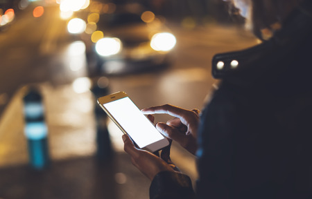 Girl pointing finger on screen smartphone on background bokeh light in night atmospheric city street, blogger hipster girl using in hands mobile phone, lights auto taxi; lifestyle online wifi internet conceptの写真素材