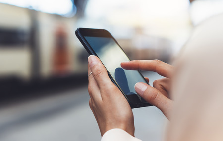 blogger hipster using in hands gadget mobile phone, woman texting message on blank screen smartphone, texting message, mockup online wifi internet concept, hipster waiting on station platform on background trainの写真素材