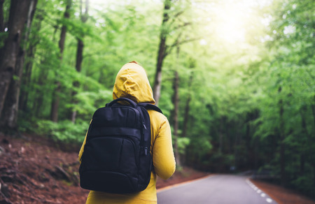 tourist traveler with backpack standing into road at summer green forest, view back girl hiker in yellow hoody looking and enjoying the breath of fresh clean air in trip, relax holiday concept, blurred backgroundの写真素材