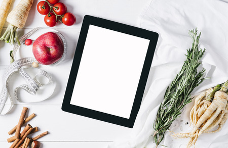 Tablet with a measuring tape, rosemary, lime, lemon, nuts, parsley root, tomatoes, cinnamon and red apples on a white wooden background top view horizontalの写真素材