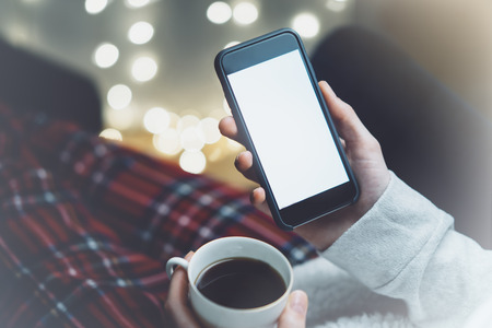 Girl pointing finger a black smartphone with a empty blank screen monitor and a cup of coffee or tea on the background bokeh light in a chair in a homely atmosphere, freelancer hipster working and using in female hands a smart phone with blank template copy space screen for your information or content, blurの写真素材