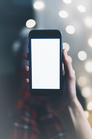 Girl pointing finger a black smartphone with a empty blank screen monitor on the background bokeh light in a homely atmosphere, freelancer hipster working at home and using in female hands smart phone with blank template copy space screen for your information or content, blurの写真素材