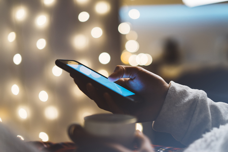 Hipster girl using mobile phone in a home atmosphere, person holding smartphone on background glow bokeh Christmas illimination, female hands texting on relax glitter xmas decoration, mockup templates, blurの写真素材