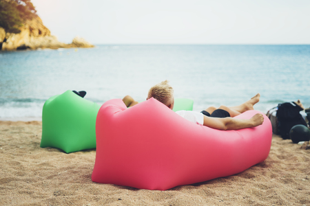 young hipster relaxing on coastline beach on inflatable lazy air pouffe sofa, person tourist enjoy sunny day on background coast sea waves, calm concept mockup, seascape perspective vacation, summer seashoreの写真素材