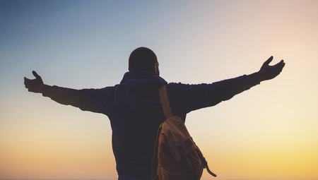 tourist traveler with backpack standing with raised hands, hiker looking on sunset to valley in trip, hipster young man enjoying peak of mountain background evening seascape ocean horizon, relax holiday conceptの写真素材
