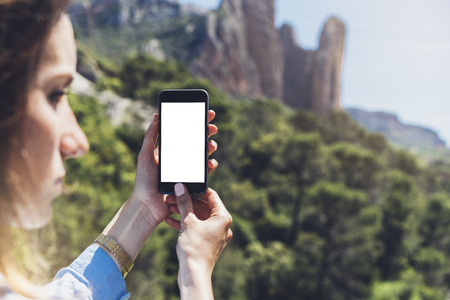 Hipster girl making photo on smartphone mobile closeup, view tourist hands using gadget phone in travel on background mountains and sky landscape; finger touch screen cellphone mockup nature, templatesの写真素材