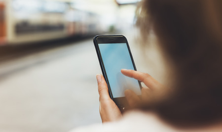 blogger hipster using in hands gadget mobile phone, woman texting message on blank screen smartphone, texting message, mockup online wifi internet concept, hipster waiting on station platform on background trainの写真素材