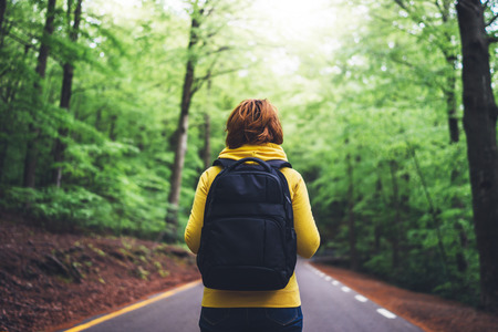 tourist traveler with backpack standing into road at summer green forest, view back girl hiker in yellow hoody looking and enjoying the breath of fresh clean air in trip, relax holiday concept, blurred backgroundの写真素材