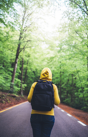 tourist traveler with backpack standing into road at summer green forest, view back girl hiker in yellow hoody looking and enjoying the breath of fresh clean air in trip, relax holiday concept, blurred backgroundの写真素材