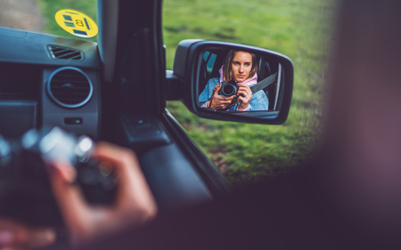 tourist girl in an open window of a car taking photography click on retro vintage photo camera, photographer looking on reflection in a mirror auto, blogger using hobby content concept, enjoy tripの写真素材