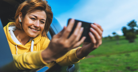smile tourist girl in an open window of a auto car taking photo selfie on mobile smart phone, person looking on camera gadget technology, blogger using content online wifi internet conceptの写真素材