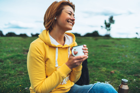 Happy smile girl holding in hands cup of hot tea on green grass in outdoors nature park, beautiful woman hipster enjoy drinking cup of coffee, lifestyle relax recreation meditation conceptの写真素材