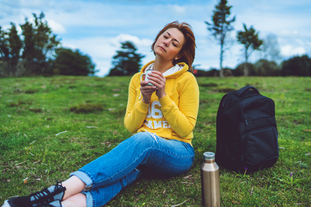 Happy girl with eyes closed holding in hands cup of hot tea on green grass in outdoors nature park, beautiful woman hipster enjoy drinking cup of coffee, lifestyle relax recreation meditation conceptの写真素材