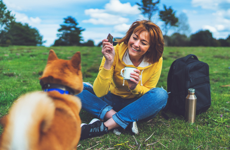 Happy girl with smile drink cup playing with red japanese dog shiba inu on green grass in the outdoors nature park, beautiful young woman hipster and dog friends, friendship lifestyle conceptの写真素材
