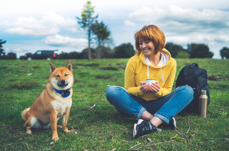 Happy smile girl holding in hands cup drink, red japanese dog shiba inu on green grass in outdoors nature park, beautiful young woman hipster and dogs friends, friendship lifestyle relax recreation meditation conceptの写真素材