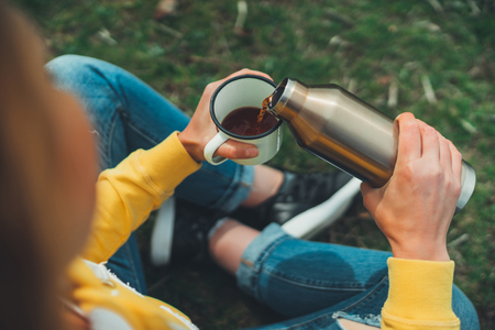 Top view girl holding in hands cup of hot tea on green grass in outdoors nature park, beautiful woman hipster enjoy drinking cup of coffee flask, lifestyle relax recreation meditation conceptの写真素材