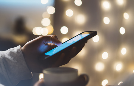 Hipster girl using mobile phone in a home atmosphere, person holding smartphone on background glow...