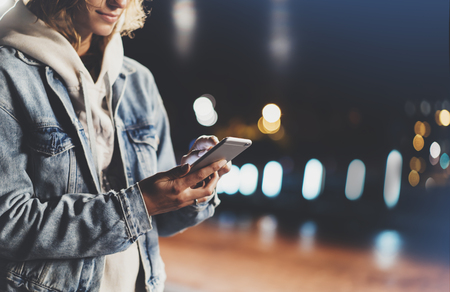 Girl pointing finger on screen smartphone on background illumination bokeh color light in night atmospheric city, hipster using in female hands and texting mobile phone, mockup glitter street, content lifestyleの写真素材