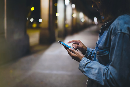Girl pointing finger on screen smartphone on background illumination glow bokeh light in night atmospheric city, hipster using in hands mobile phone, mockup glitter street in Barcelona, online internet conceptの写真素材