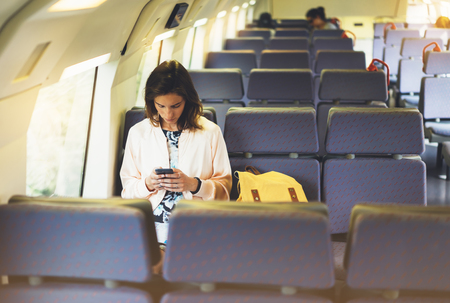 Enjoying travel. Young pretty woman traveling by train sitting near the window using smartphone and looking map. Tourist texting online message and plan route of railway, railroad transport conceptの写真素材