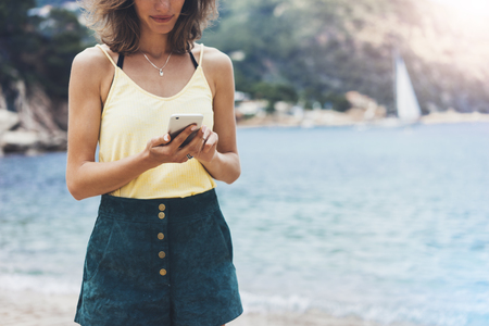 Hipster girl hold on smart phone gadget in sand coastline, mock up blank screen. Traveler using in female hand mobile on background beach seascape horizon. Tourist look on blue sun ocean, summer lifestyleの写真素材
