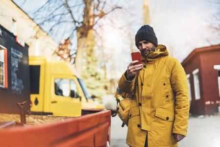 Hipster with yellow backpack, jacket, cap, coffee of thermo cup holding in the hands smartphone, freelance using gadget mobile in autumn street outdoor, tourist man typing message, online processの写真素材