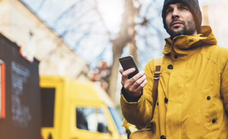 Hipster with yellow backpack, jacket, cap, coffee of thermo cup holding in the hands smartphone, freelance using gadget mobile in autumn street outdoor, tourist man typing message, online processの写真素材