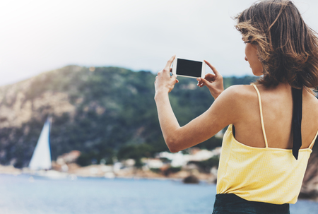 Hipster girl photograph on smart phone gadget in sand coastline, mock up of blank screen. Traveler hold and using in female hand mobile on background seascape horizon. Tourist and blue sun ocean, lifestyleの写真素材