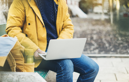 Hipster with yellow backpack, coat, drink coffee of thermo cup, freelance using computer open laptop in autumn street outdoor, tourist man typing on keyboard, traveler connect wifi internet, process in workspaceの写真素材