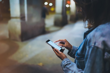 Girl pointing finger on screen smartphone on background illumination glow bokeh light in night atmospheric city, hipster using in hands mobile phone, mockup glitter street in Barcelona, online internet conceptの写真素材
