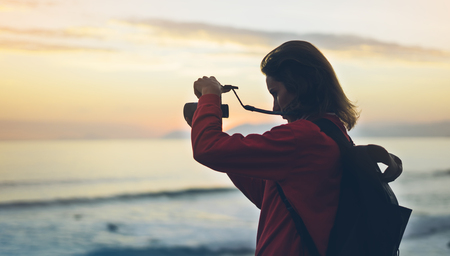 Hipster hiker tourist with backpack taking photo of amazing seascape sunset on camera on background blue sea, photographer enjoying ocean horizon, blurred panoramic sunrise blank mockup, traveler relax holiday concept, sunlight view in trip vacation northern spainの写真素材