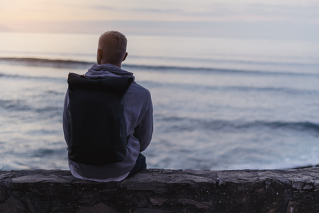 Hipster hiker tourist with backpack looking of amazing seascape sunset on background blue sea, guy enjoying ocean horizon, blurred panoramic sunrise blank mockup, traveler relax holiday concept, sunlight view in trip vacation northern spainの写真素材