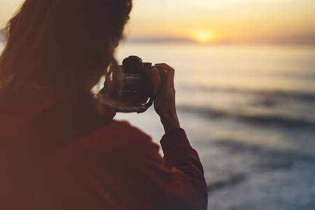 Hipster hiker tourist with backpack taking photo of amazing seascape sunset on camera on background blue sea, photographer enjoying ocean horizon, blurred panoramic sunrise blank mockup, traveler relax holiday concept, sunlight view in trip vacation northern spainの写真素材