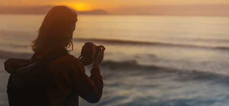 Hipster hiker tourist with backpack taking photo of amazing seascape sunset on camera on background blue sea, photographer enjoying ocean horizon, blurred panoramic sunrise blank mockup, traveler relax holiday concept, sunlight view in trip vacation northern spainの写真素材