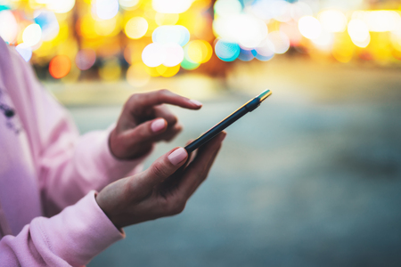 Person pointing finger on screen smartphone on defocus background bokeh light in evening street, hipster girl using in hands mobile phone gadget in night atmospheric city illumination, online wifi internet conceptの写真素材