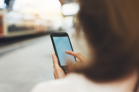 blogger hipster using in hands gadget mobile phone, woman texting message on blank screen smartphone, texting message, mockup online wifi internet concept, hipster waiting on station platform on background trainの写真素材