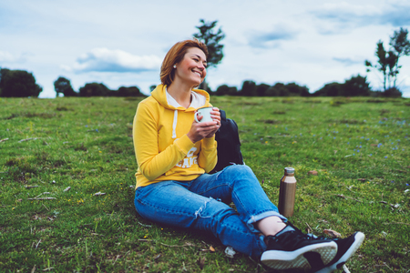 Happy smile girl holding in hands cup of hot tea on green grass in outdoors nature park, beautiful young woman hipster enjoy drinking cup of coffee, lifestyle relax recreation meditation conceptの写真素材