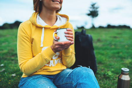 Happy smile girl holding in hands cup of hot tea on green grass in outdoors nature park, beautiful young woman hipster enjoy drinking cup of coffee, lifestyle relax recreation meditation conceptの写真素材