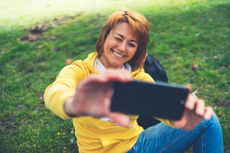 tourist girl on background green grass taking photo selfie on mobile smart phone, person looking on camera gadget technology, blogger using content online wifi internet lifestyle conceptの写真素材